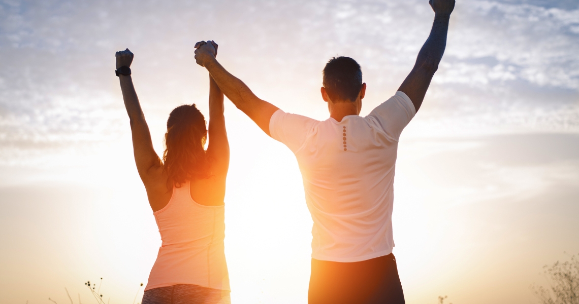 A man and woman holding hands above their head with the sunset in front of them.