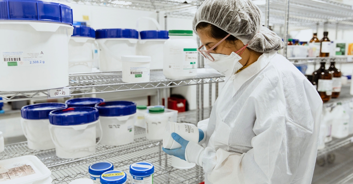 A person in full protective gear reading a white container with a metal shelf of other containers behind them.