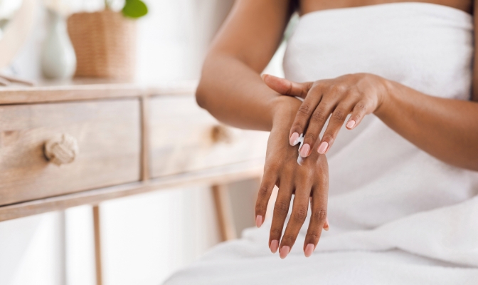 An African American woman applying lotion to her hands in a white bath towel seated at a desk.