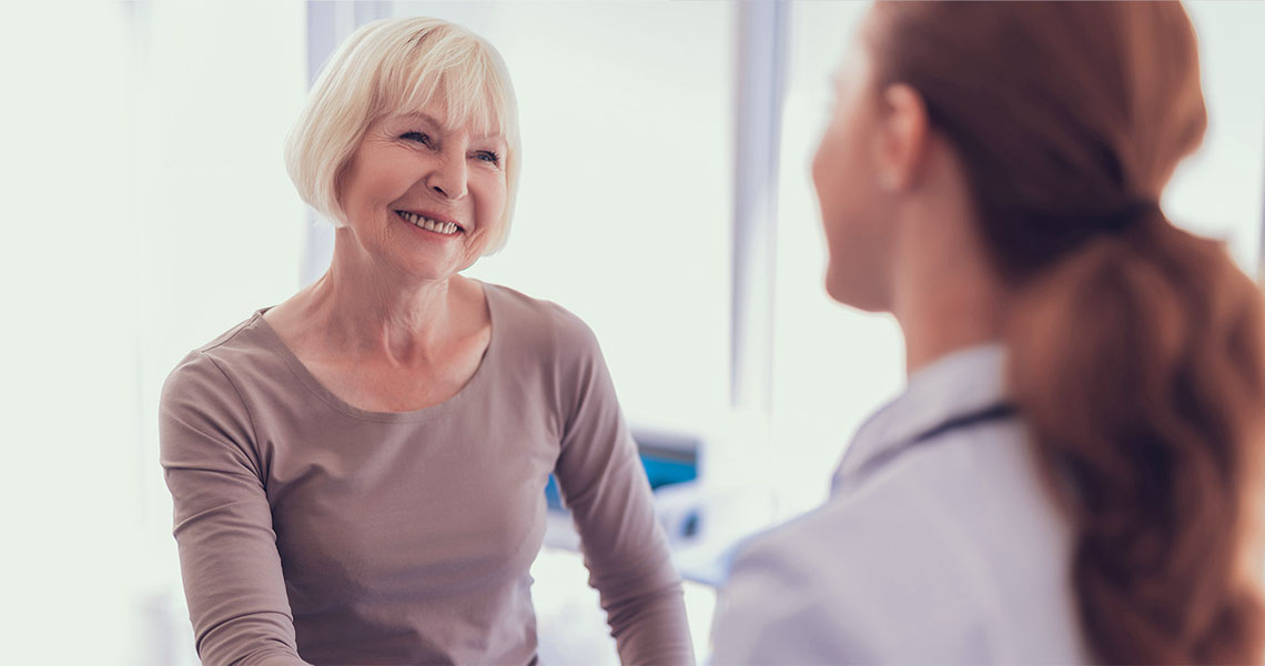 An older woman in a tan long sleeved shirt smiling while speaking to a provider with a plain gray green background.