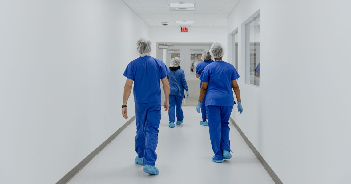 A view from the back of three people in navy blue scrubs and protective gear walking down a white hallway.