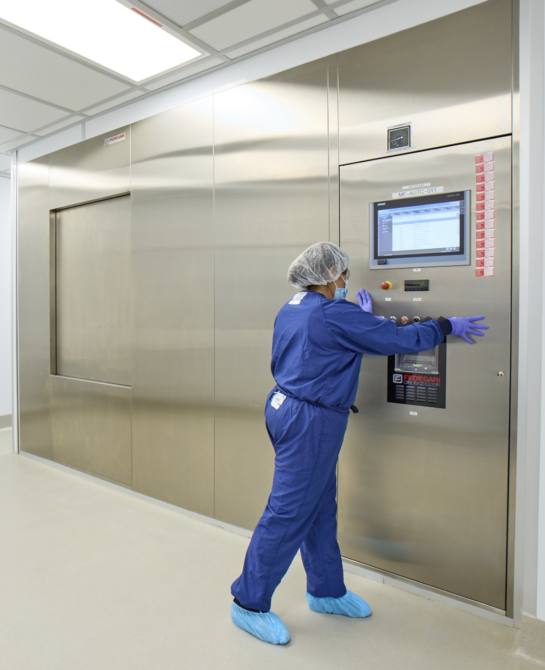 A worker in blue scrubs, a mask, and hairnet touching a large machine door.